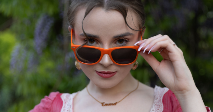 Emma dressed in a vivid pink regency gown, with neon orange sunglasses and a cheeky expression